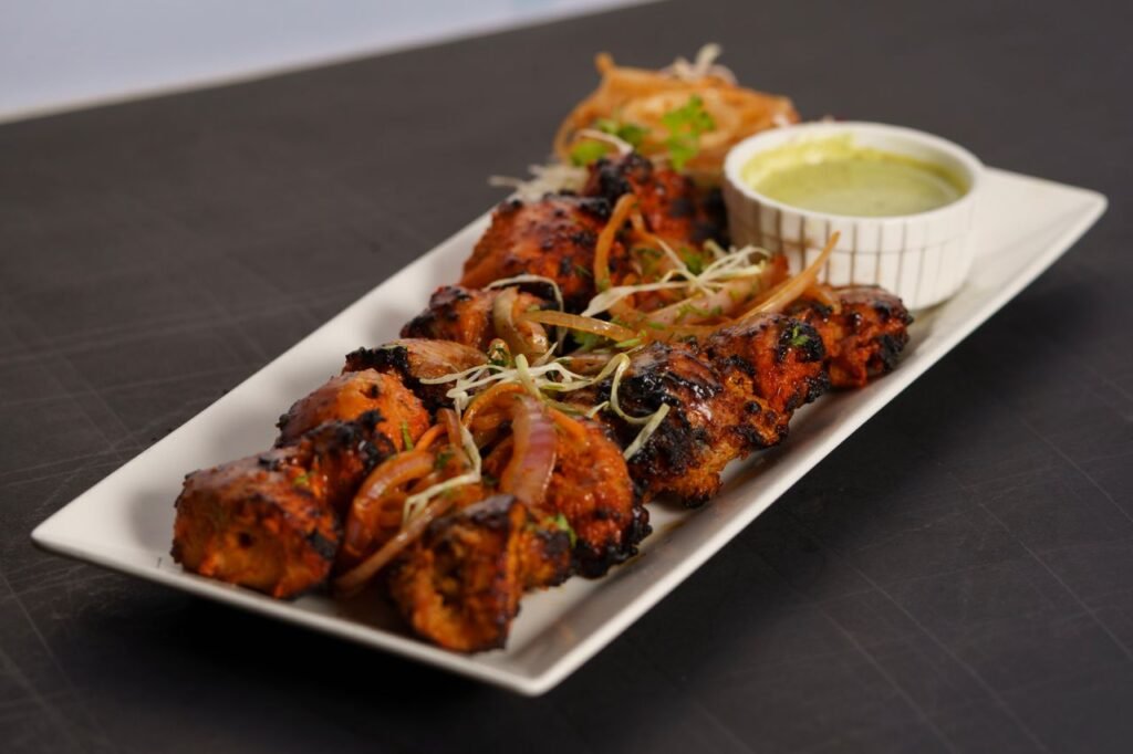 HOME A flavorful chicken tikka dish served with a side of green chutney and garnishes on a white platter.