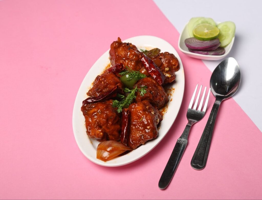HOME Delicious Chicken Manchurian garnished with herbs, served on a pink background with cutlery.